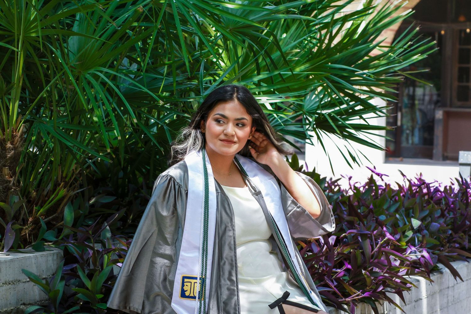 A student in a gray graduation gown and white top poses near a lush palm frond at her senior photography session at The Pearl in San Antonio