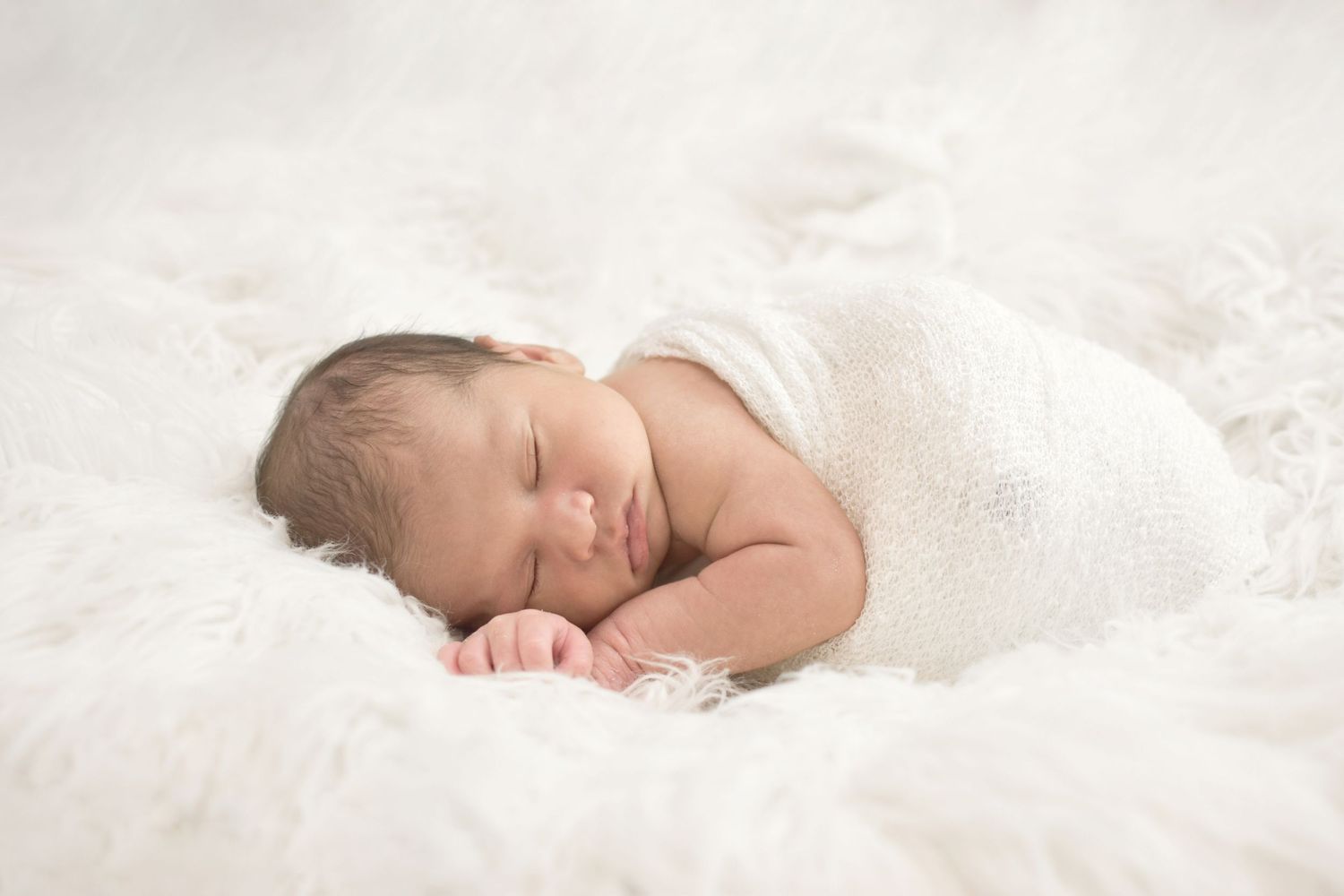 Peaceful newborn baby sleeping wrapped in white on a soft fluffy white blanket in a serene studio setting.