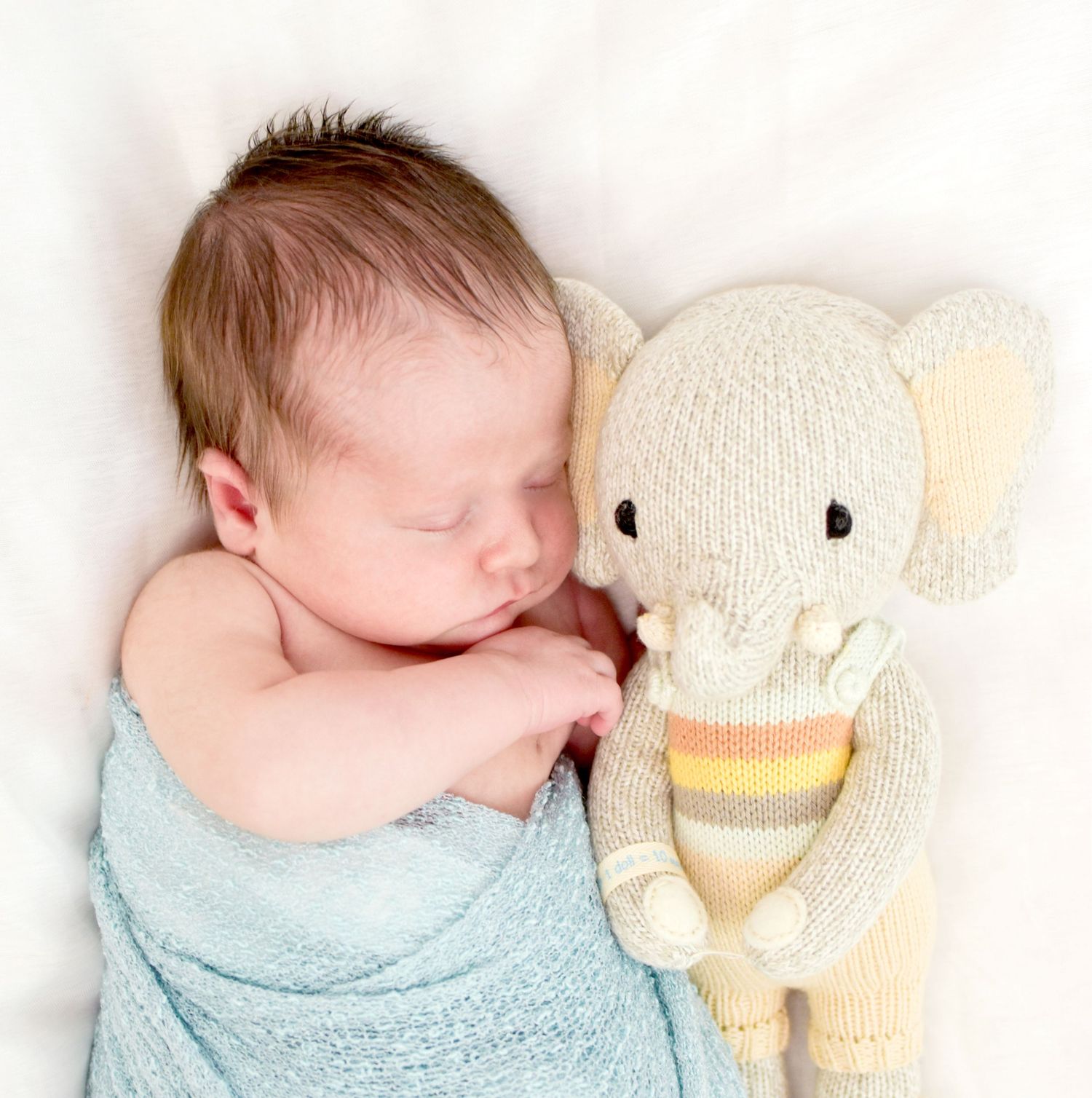 Sleeping newborn baby wrapped in blue blanket cuddled next to a knitted elephant stuffed animal toy.