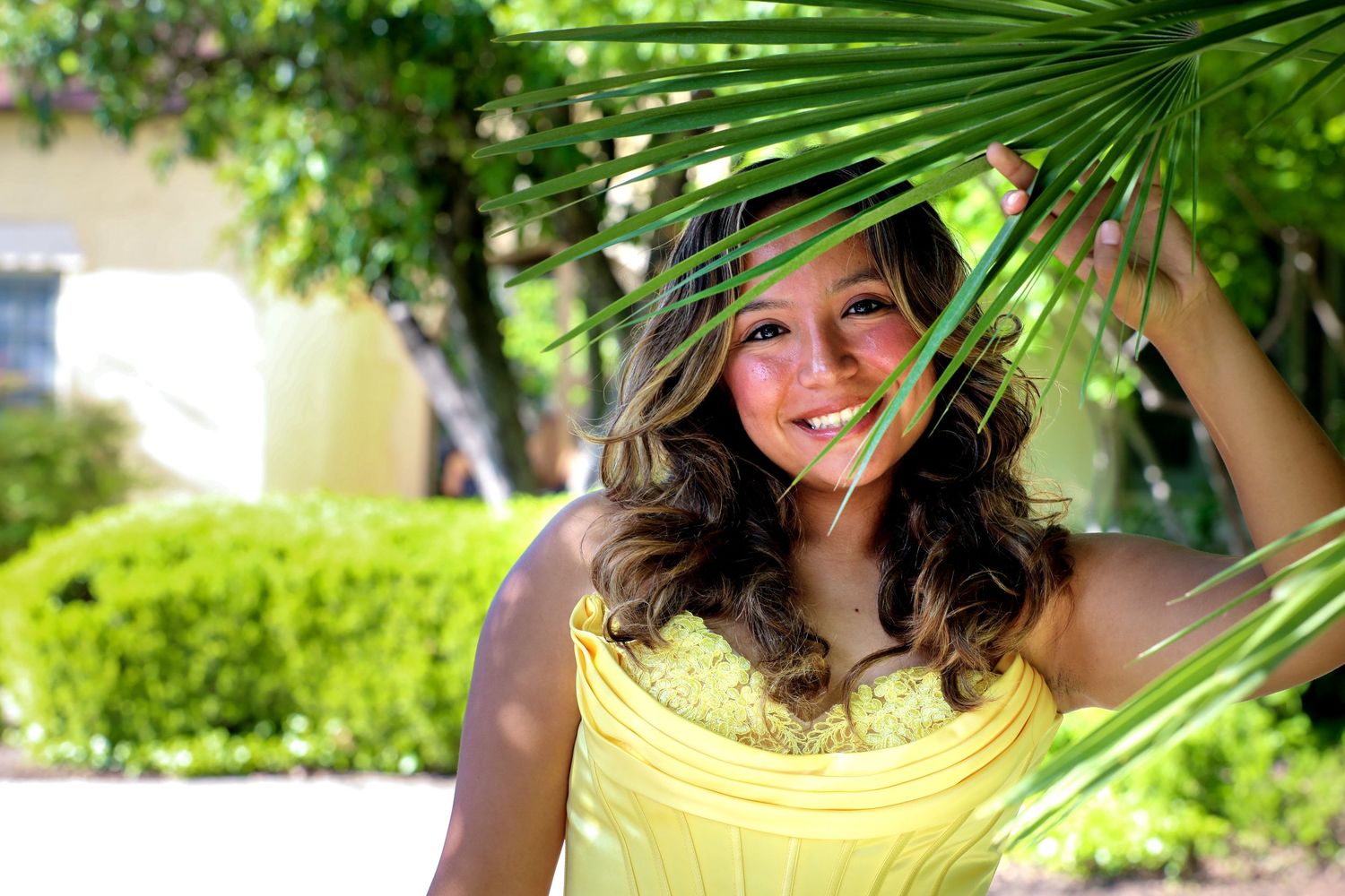 Young girl in yellow dress poses with palm fronds at Landa Library in San Antonio, Texas during her outdoor senior photo session.