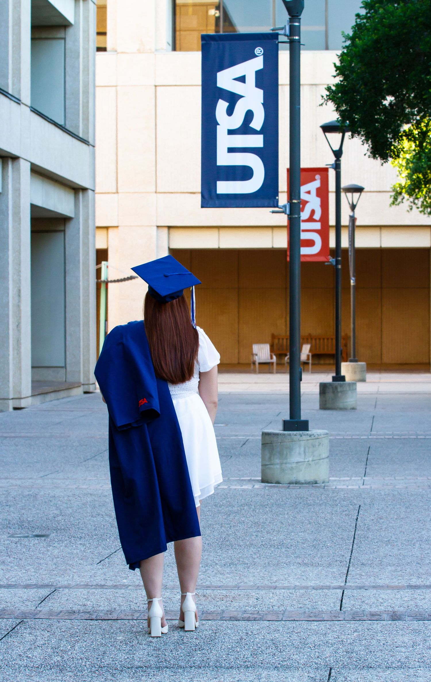 A UTSA student walking in navy blue graduation regalia on a concrete plaza with flags at university during her graduation photo  session.