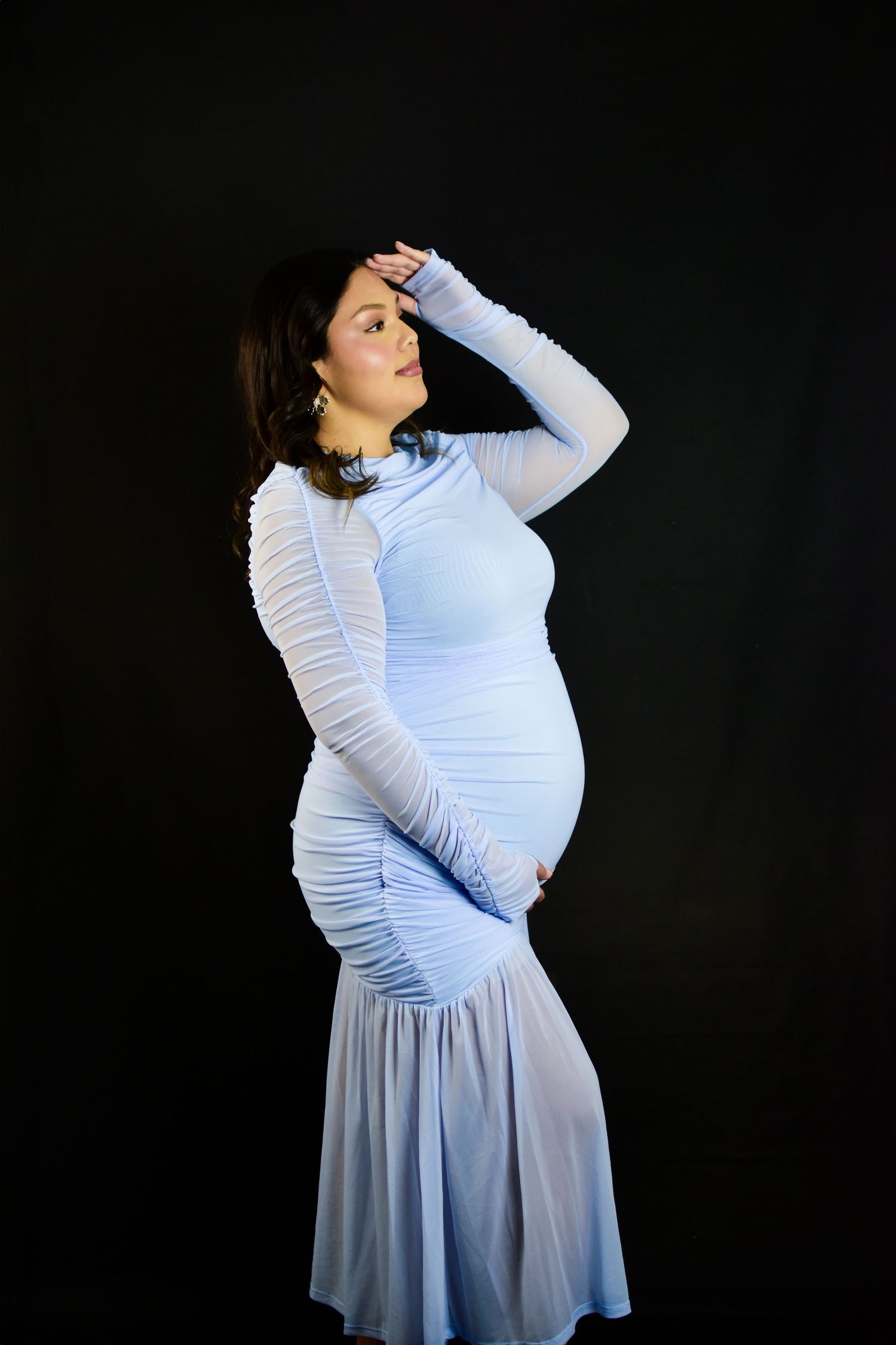 Pregnant woman in elegant light blue form-fitting dress posing against black background, hand raised to forehead.