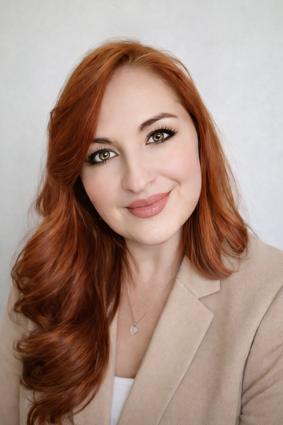 Professional headshot of a smiling woman with long red hair and green eyes wearing a beige blazer.