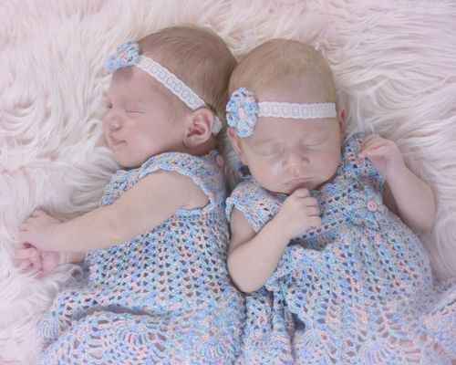 Two newborn twin babies sleeping peacefully in matching blue crochet dresses and floral headbands on a soft pink blanket.