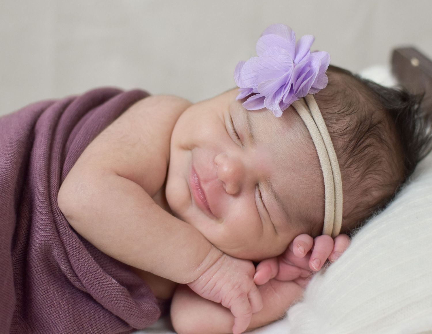 Smiling newborn baby girl sleeping peacefully wrapped in purple, wearing a lavender flower headband.