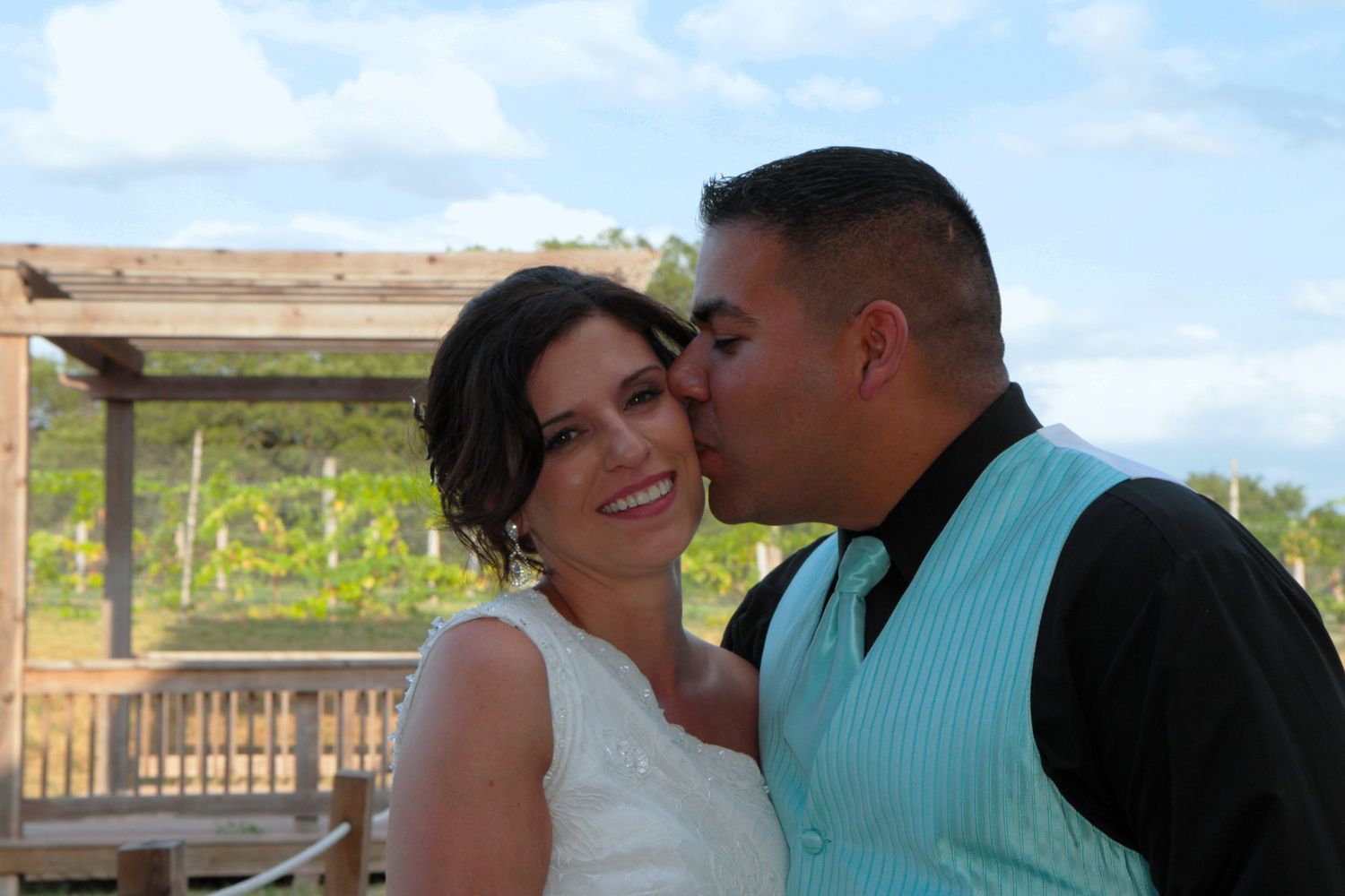 Groom in teal vest kisses bride on cheek outdoors at vineyard wedding venue under blue sky.