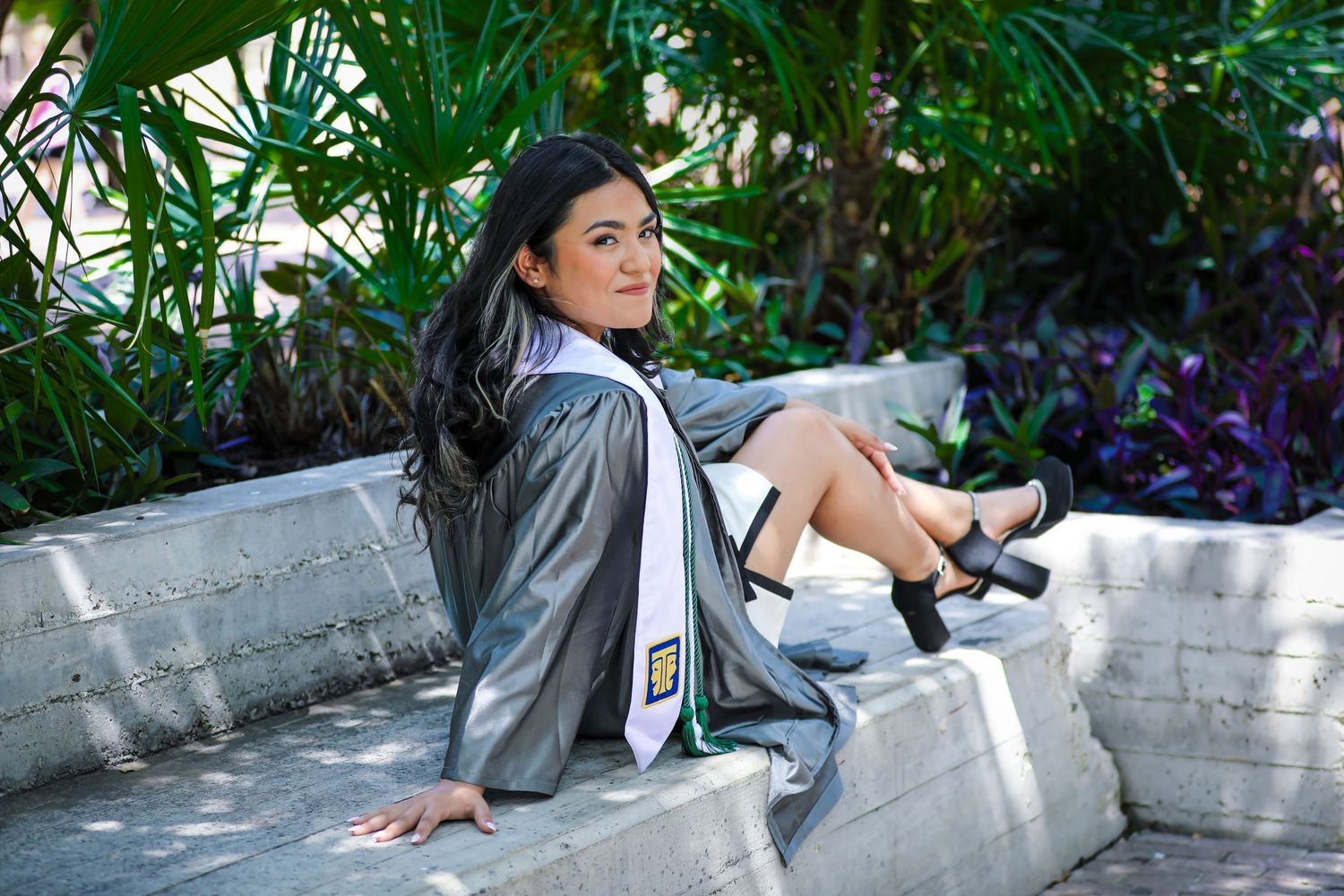 Young woman in gray graduation gown and sash sits on stone bench outdoors, smiling over her shoulder.