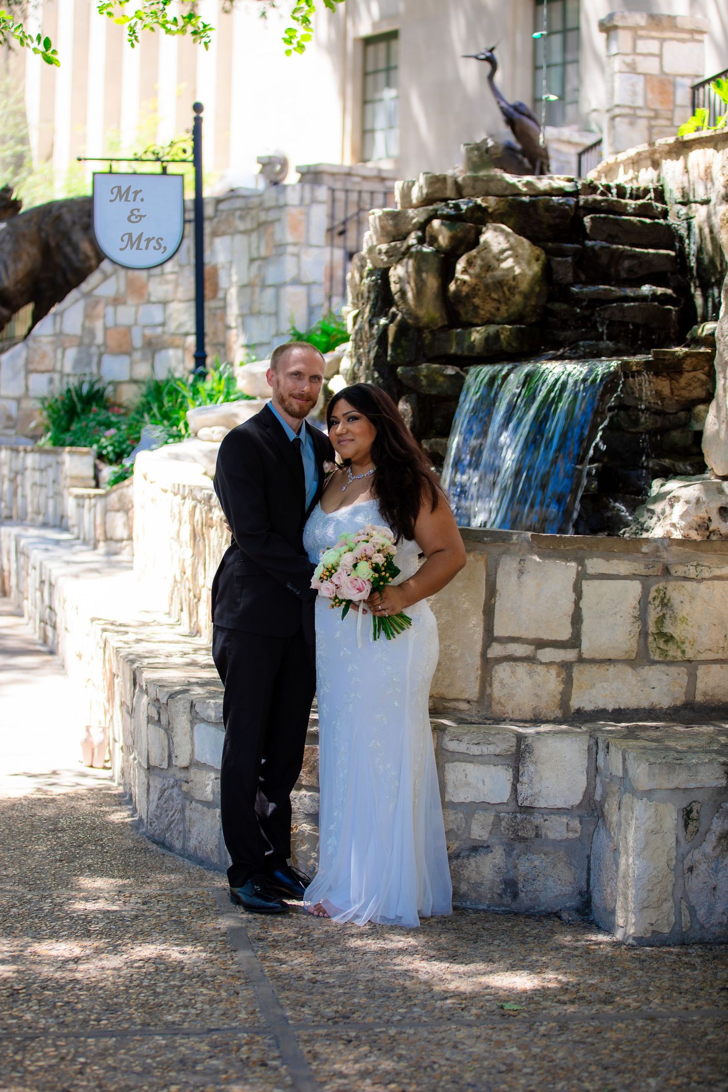 Newlyweds posing against a rustic stone wall with a waterfall on Riverwalk, San Antonio wedding photography