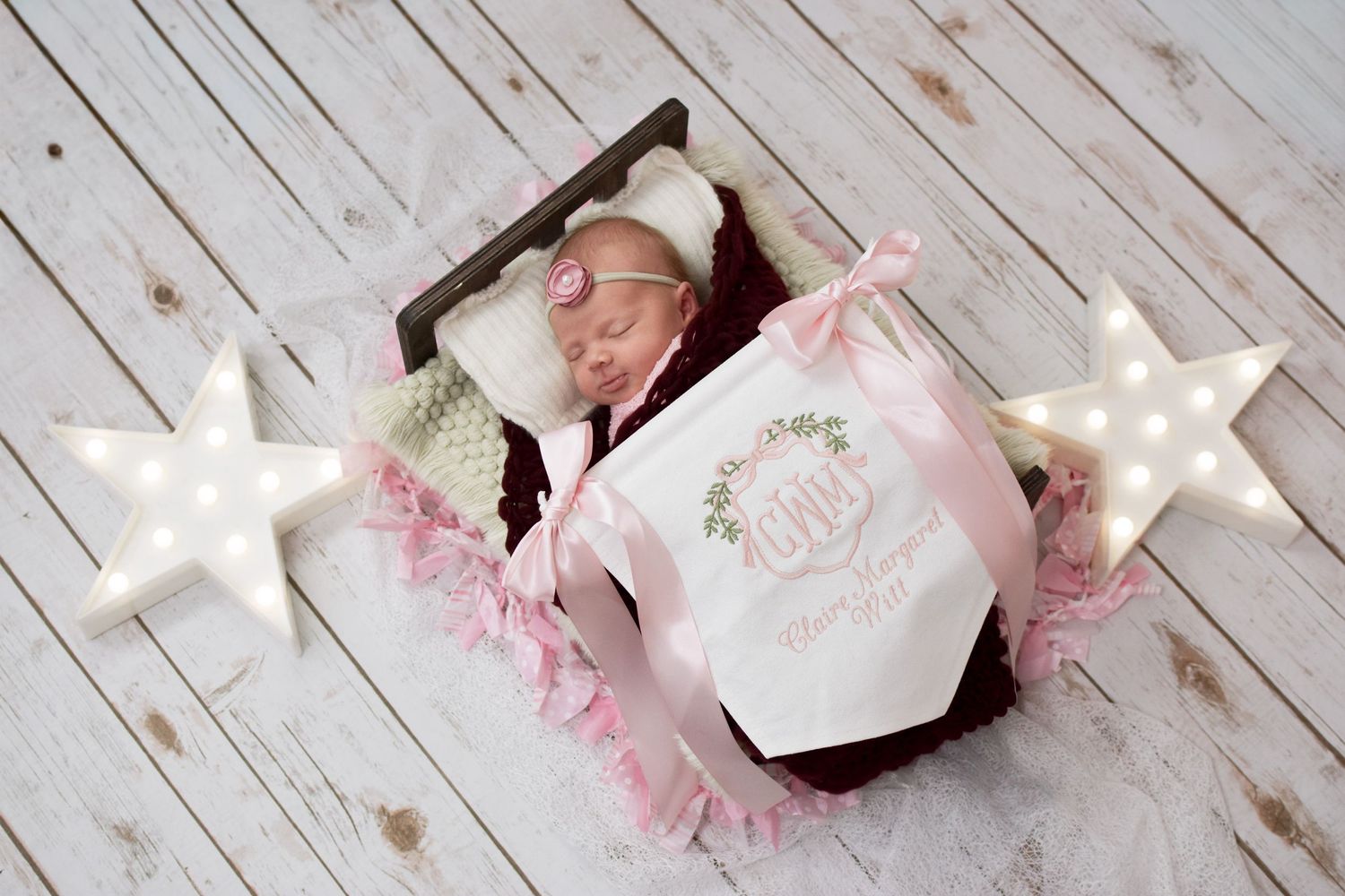 Newborn baby girl sleeping in wooden box with pink ribbon, marquee stars, and personalized monogram blanket.