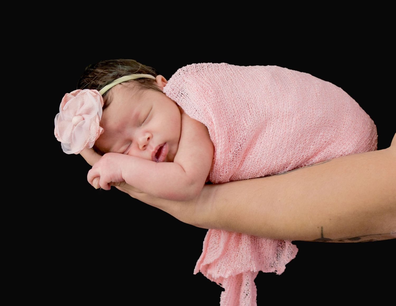 Newborn baby girl sleeping peacefully in pink wrap and floral headband, cradled in adult hands on black background.