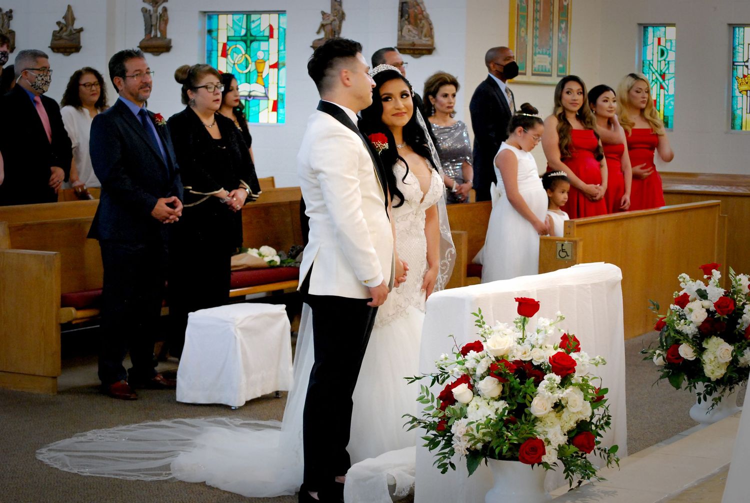 Bride and groom stand at church altar during wedding ceremony with guests in pews and red rose floral arrangements.