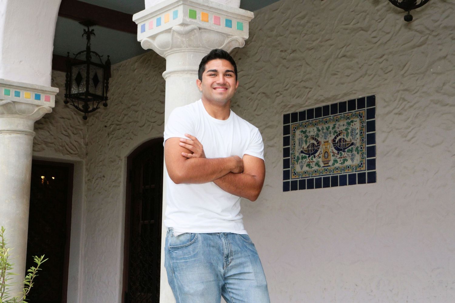 Smiling man in white t-shirt and jeans leaning against decorative column of Spanish-style building.
