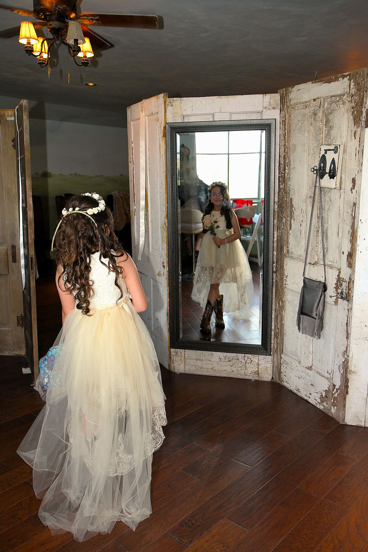 A bride's daughter in a flowing white gown walks through a rustic room with wooden floors and distressed walls. San Antonio wedding photos
