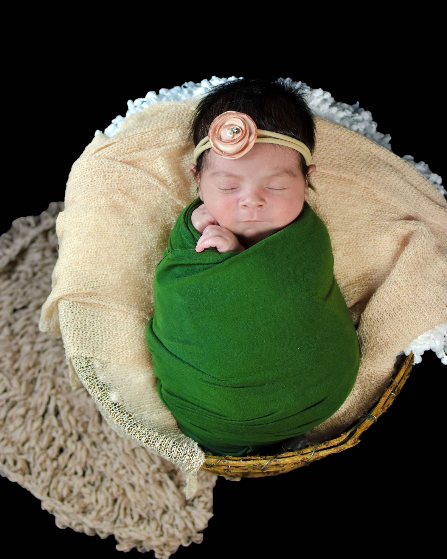 Newborn baby swaddled in green wrap sleeping peacefully in a wicker basket with a pink flower headband.