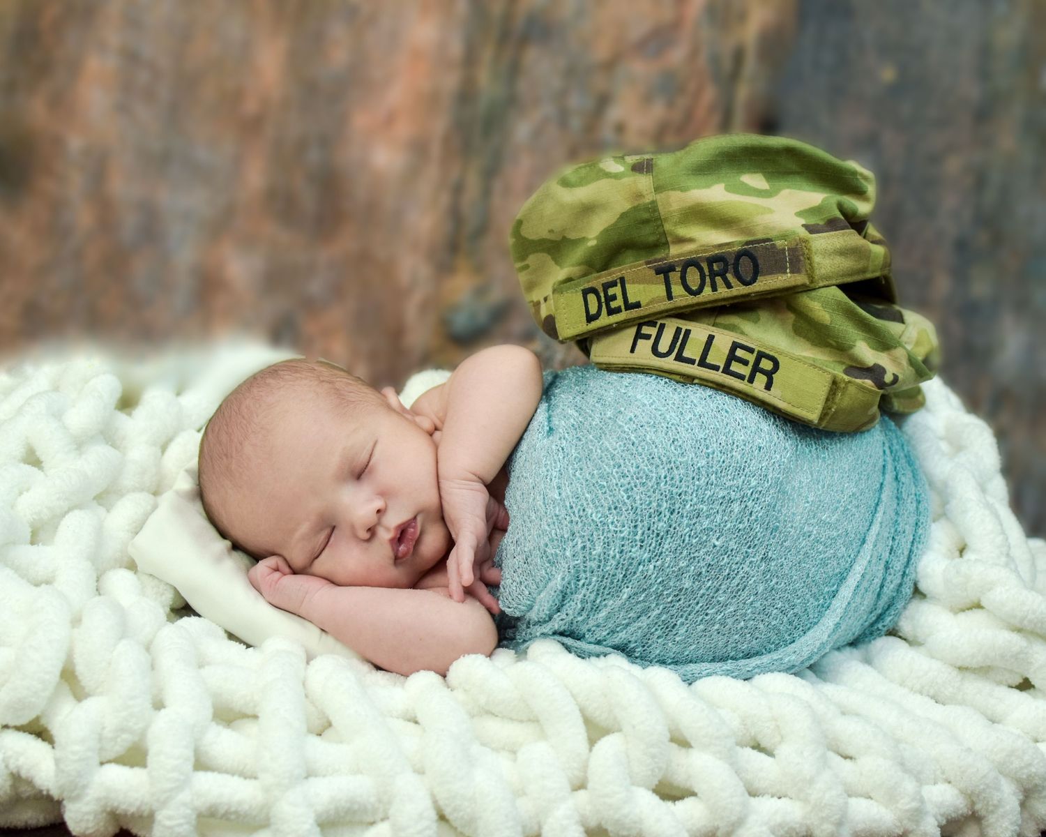 Sleeping newborn baby wrapped in blue swaddle on white knit blanket wearing military camouflage hat with name tapes.