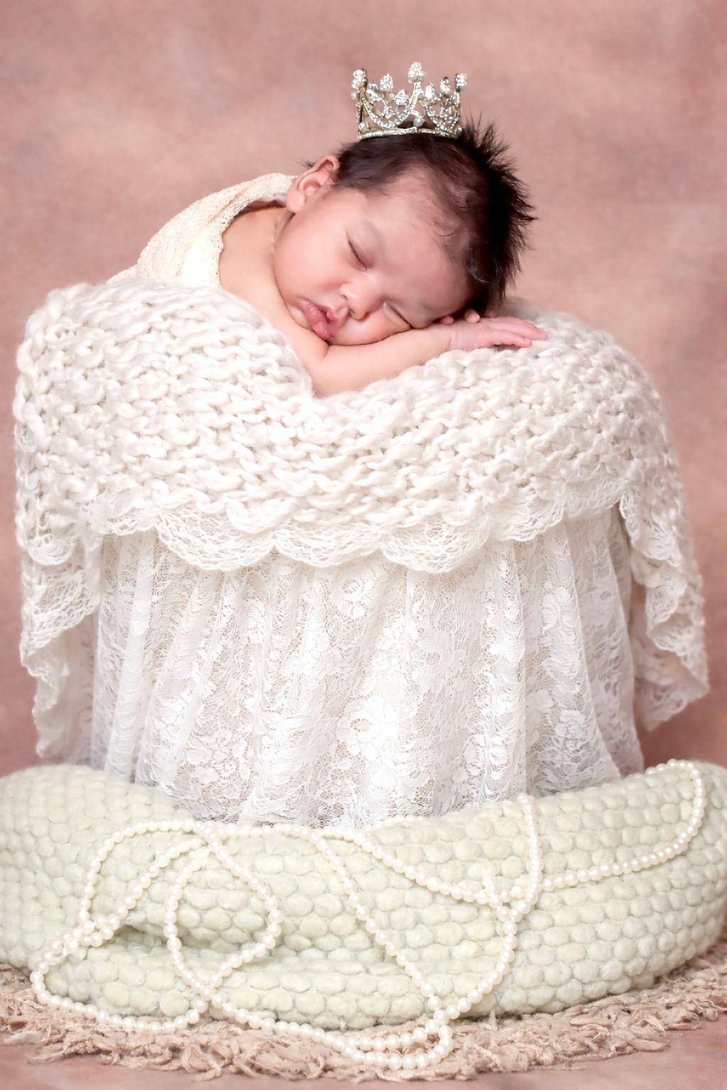 Sleeping newborn baby wearing a tiny silver crown nestled in a white lace basket with pearl beads.