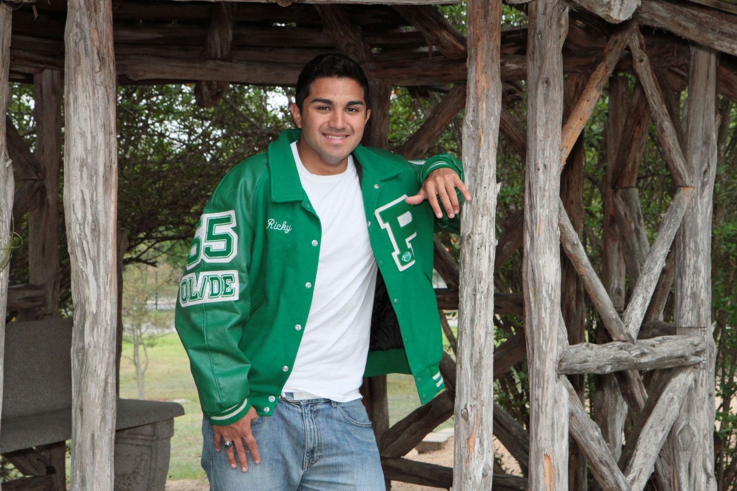 A student wearing his green varsity jacket poses under a wooden structure outdoors at the McNay Museum during his photography session.