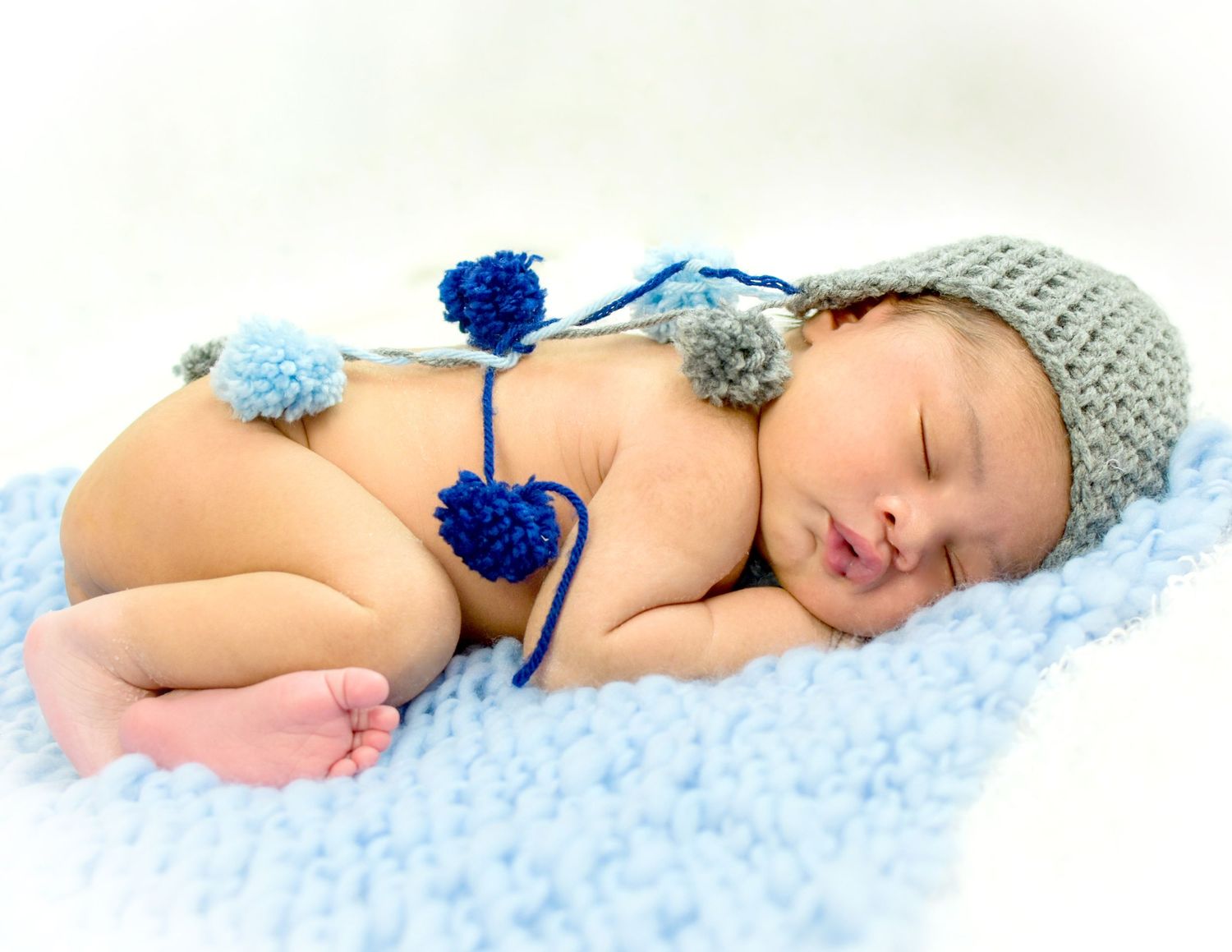 Sleeping newborn baby wearing a gray knit hat with blue pom-poms, resting on a soft blue blanket.