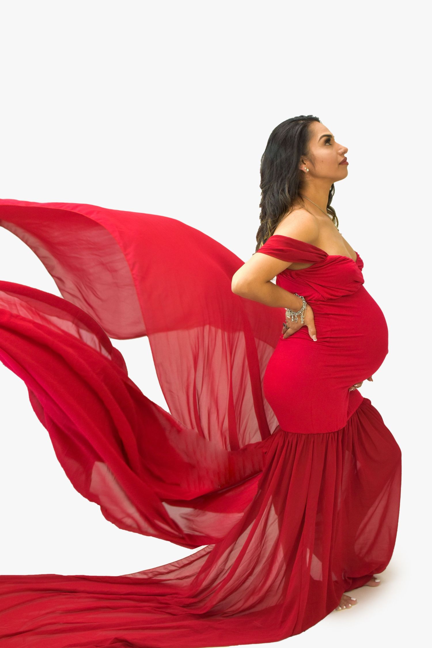 Pregnant woman in flowing red off-shoulder gown with fabric billowing dramatically against white background.