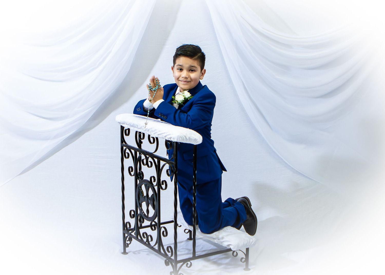 Young boy in royal blue suit kneeling at ornate white podium, formal portrait with white drape background.
