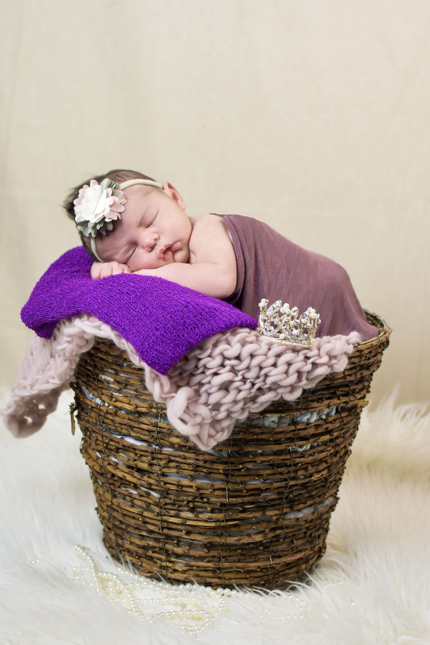 Sleeping newborn baby girl in a wicker basket with purple blankets and a floral headband.