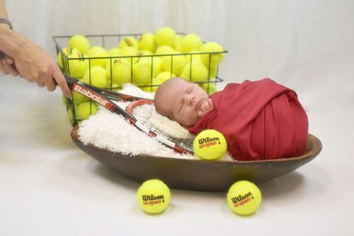 Newborn baby swaddled in red wrap sleeping in a bowl surrounded by Wilson tennis balls and a racket.