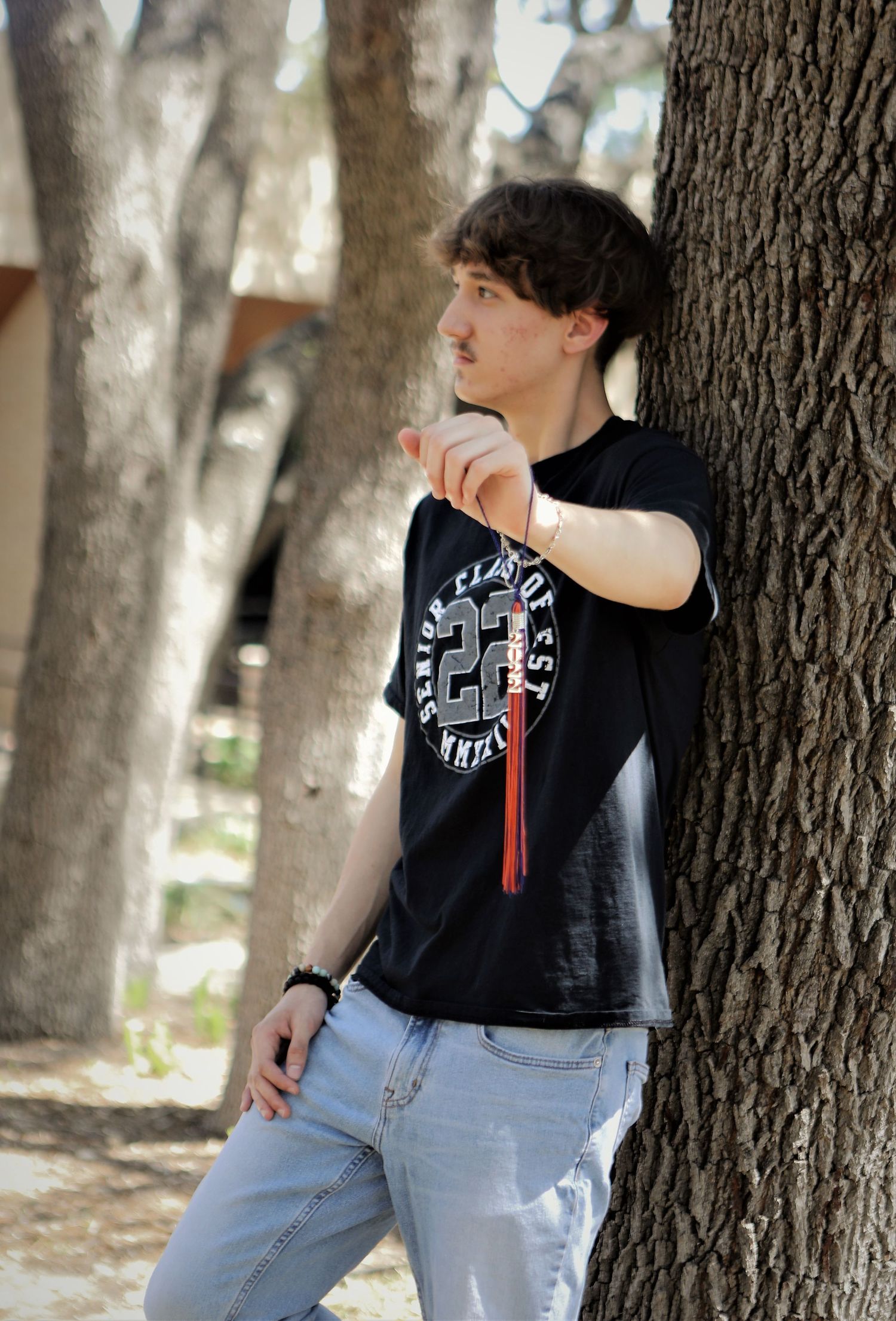 A student in a black senior t-shirt and light jeans leans against a tree trunk in San Antonio at his senior photo session hold his tassel.