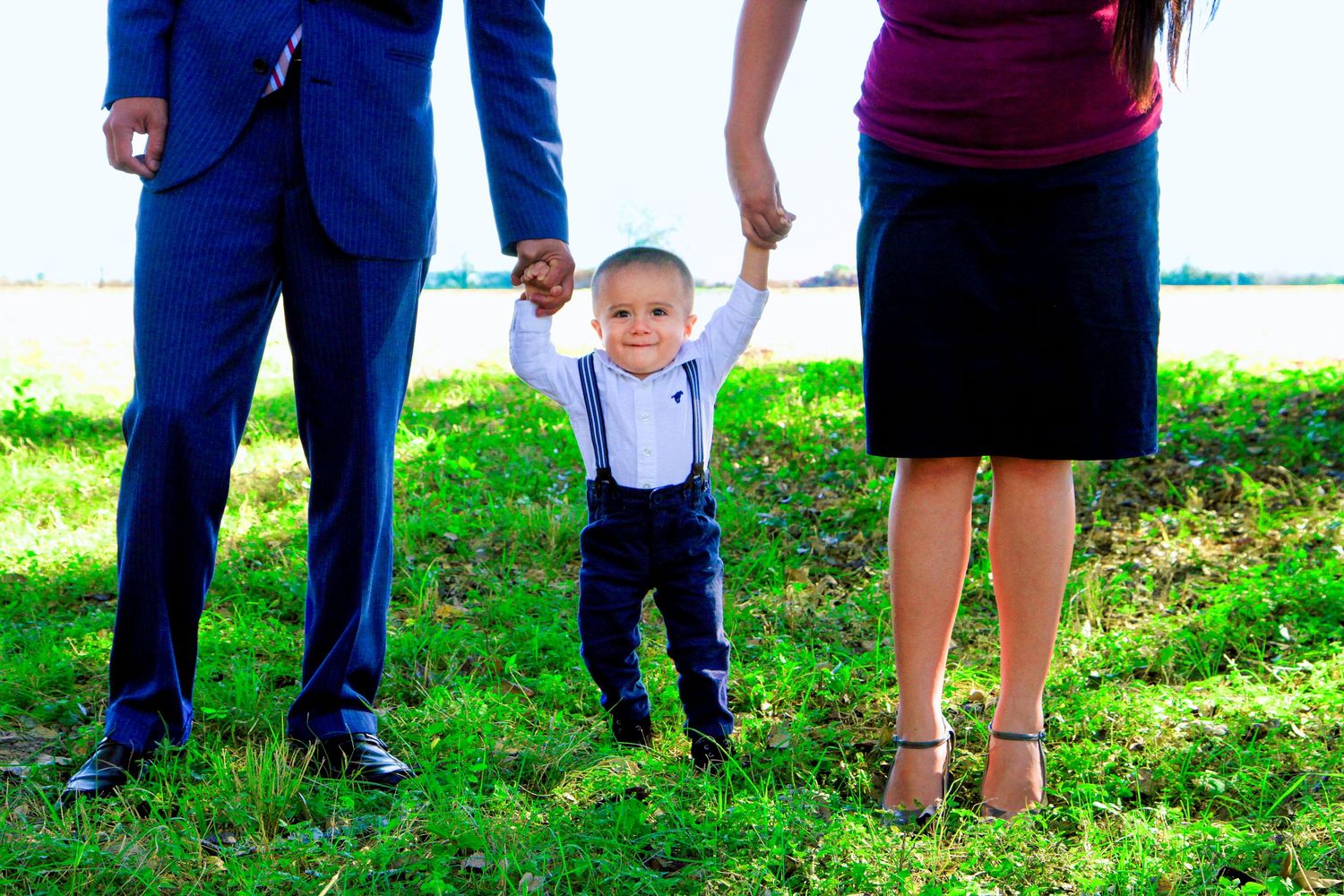 A toddler boy in suspenders holds hands with his parents outdoors on green grass near a waterfront.