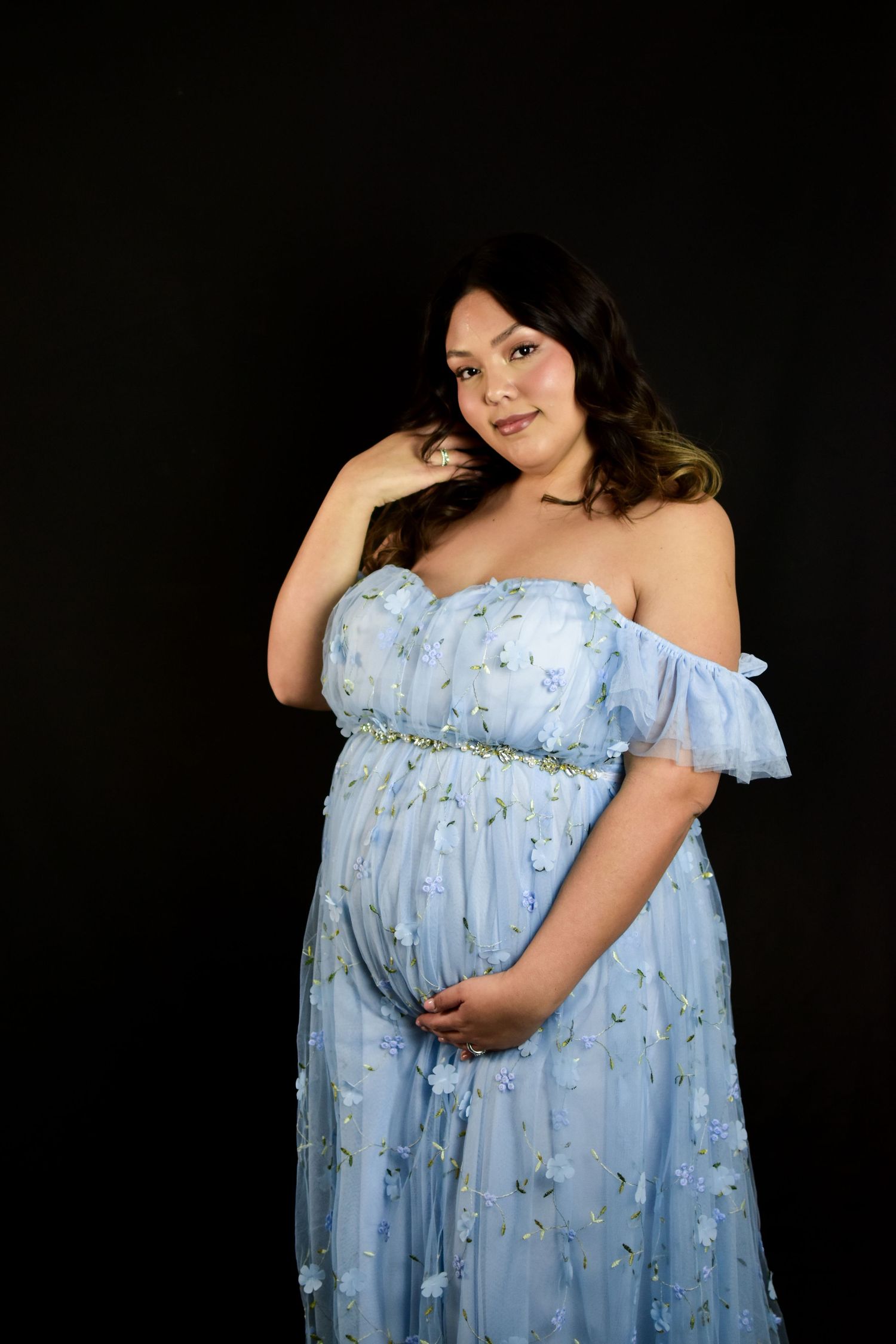 Pregnant woman in a blue floral off-shoulder gown posing gracefully against a dark background.