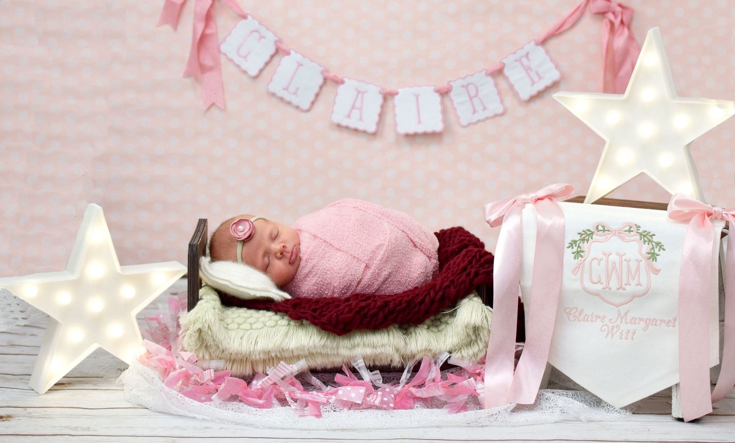 Newborn baby girl sleeping in a tiny bed wrapped in pink, surrounded by star lights and a monogram gift box.