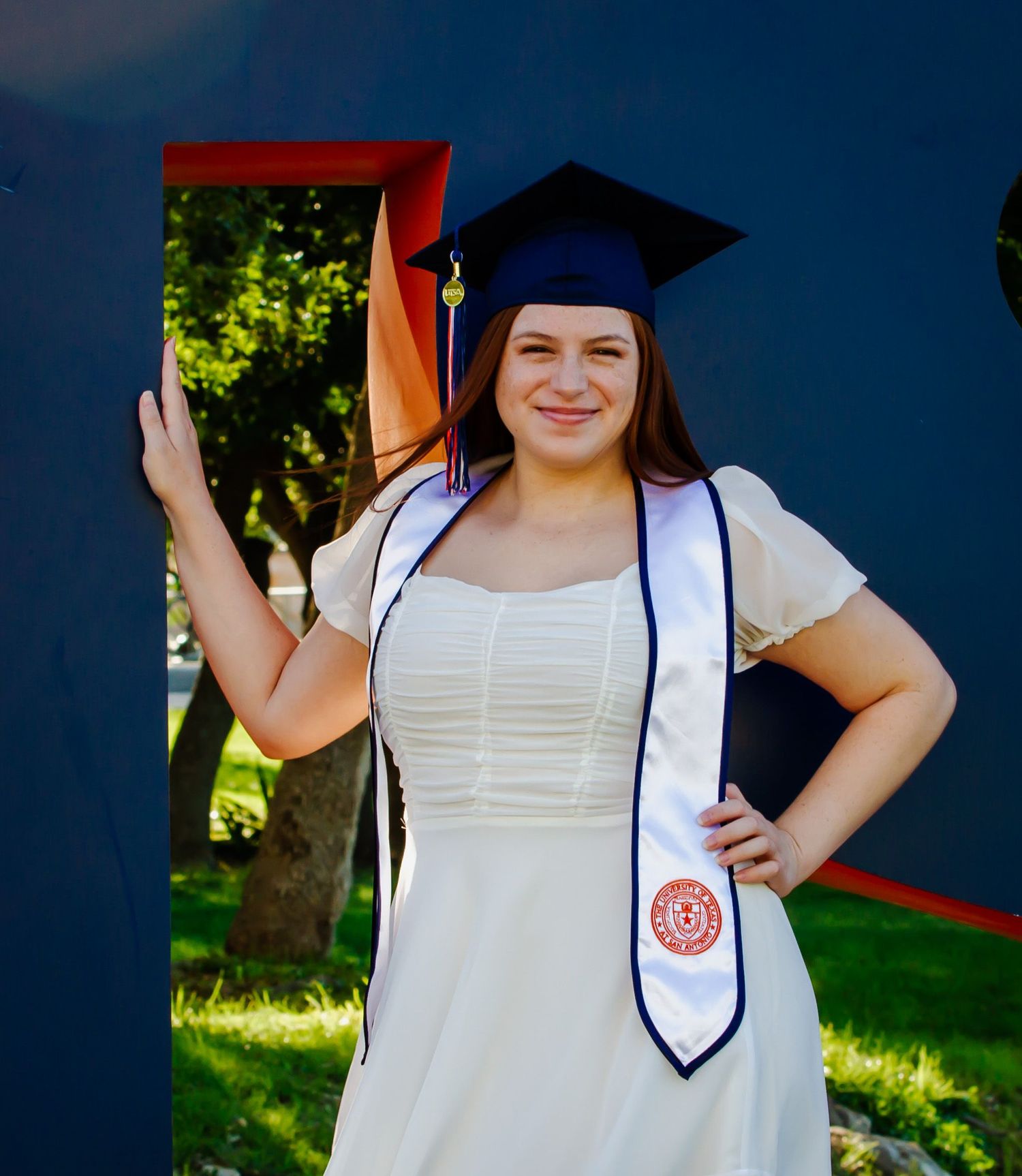 Graduate in white dress and UTSA graduation cap wearing ceremonial sash during outdoor graduation/senior photo session in San Antonio, Tx.