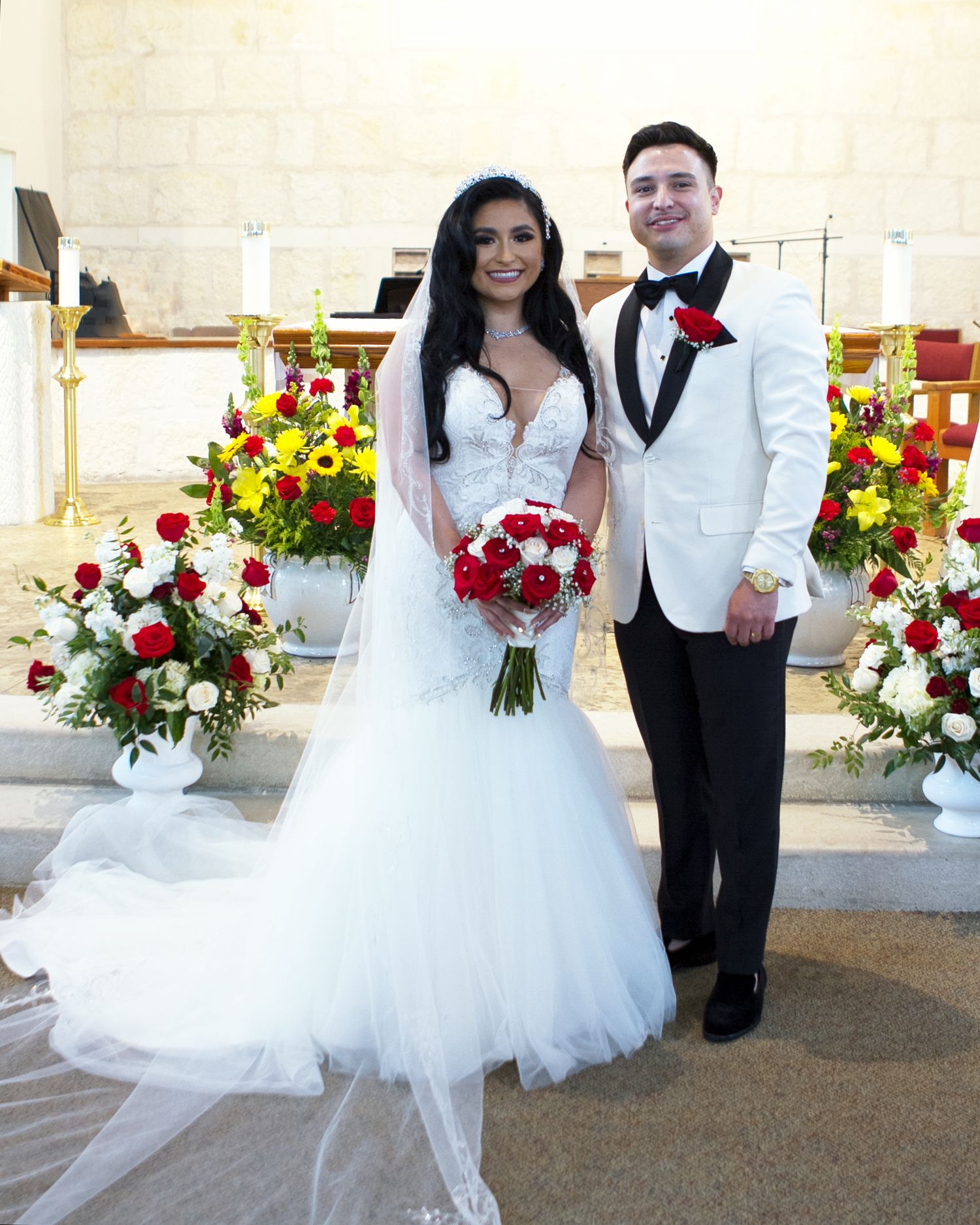 Bride and groom posing at church altar with red and yellow floral arrangements, bride in mermaid gown holding red rose bouquet.