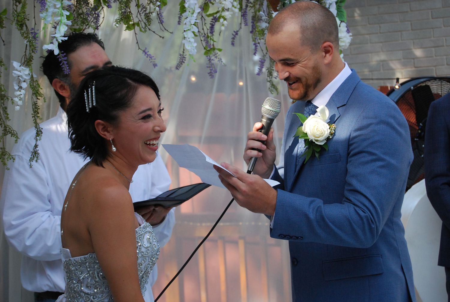 Bride and groom laughing during wedding ceremony vows, groom in blue suit with white boutonniere holding microphone.