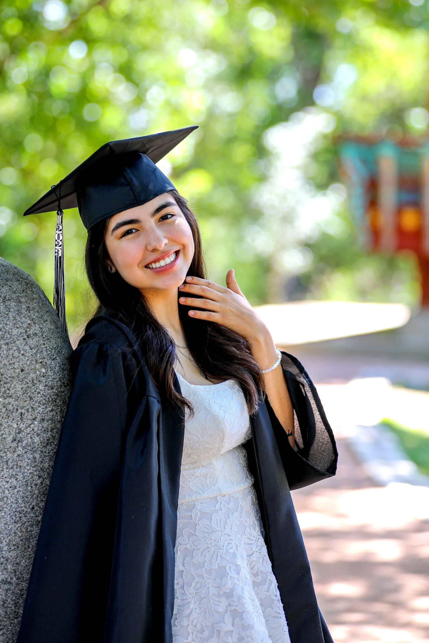Smiling young woman in black graduation cap and gown posing outdoors in a lush green park setting.