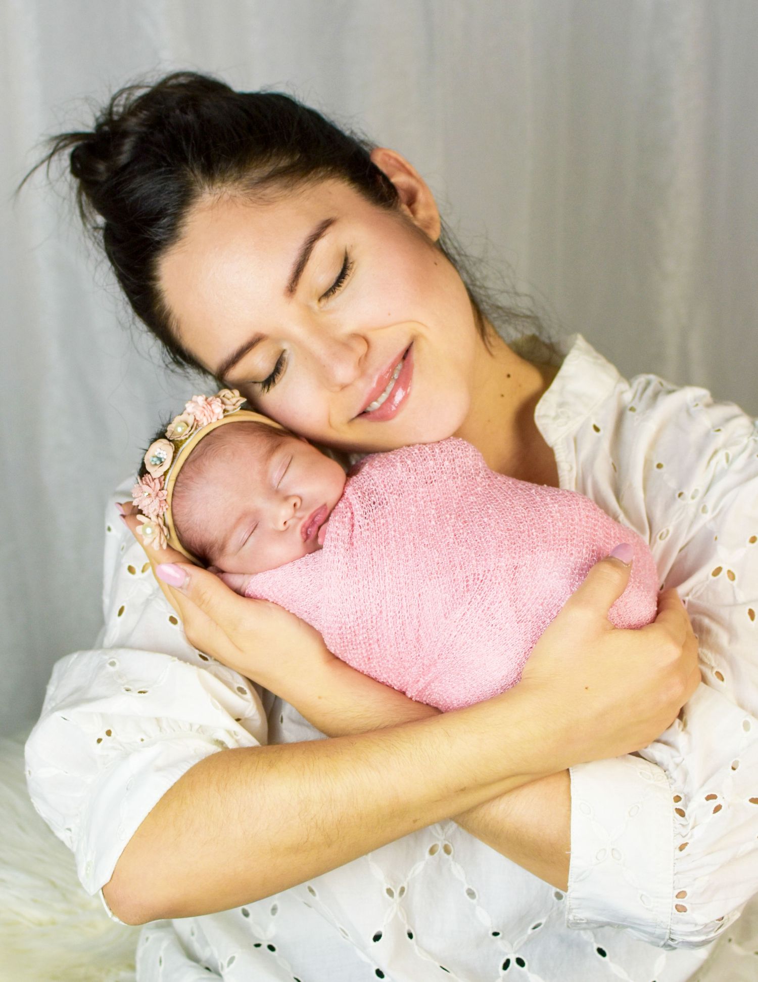 A smiling mother with closed eyes tenderly holds her sleeping newborn baby wrapped in a pink swaddle with a floral headband.