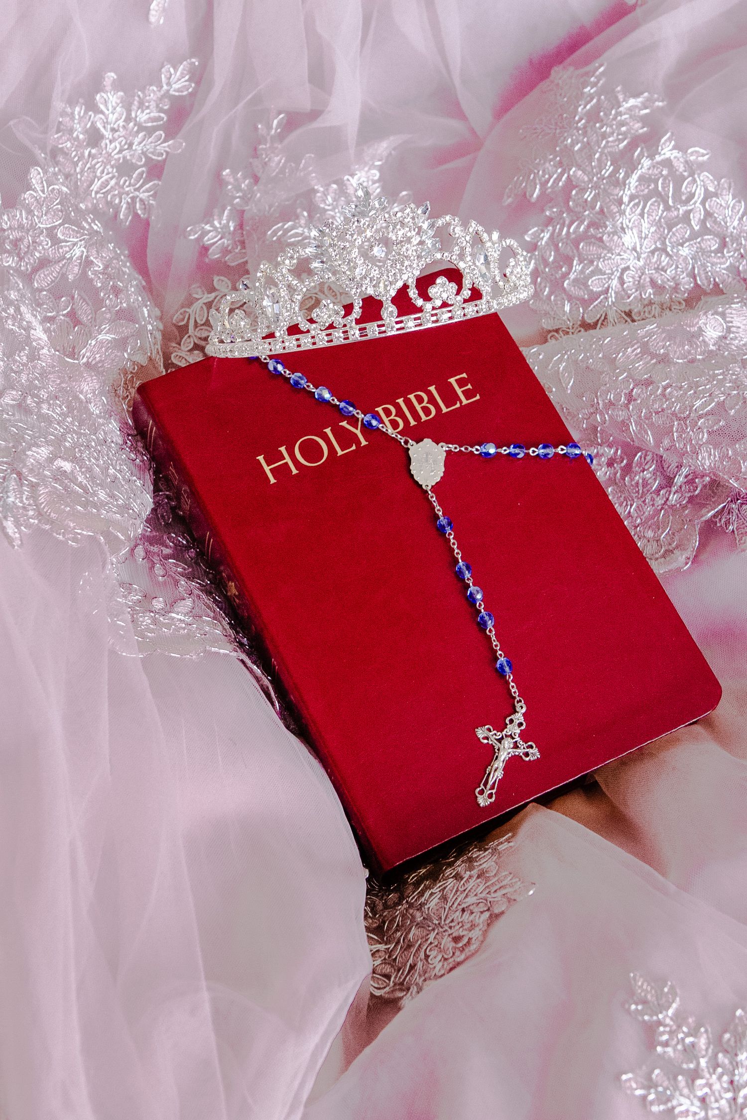 A red Holy Bible with a delicate blue beaded rosary with cross draped across it on pink lace during a quinceanera photography session.