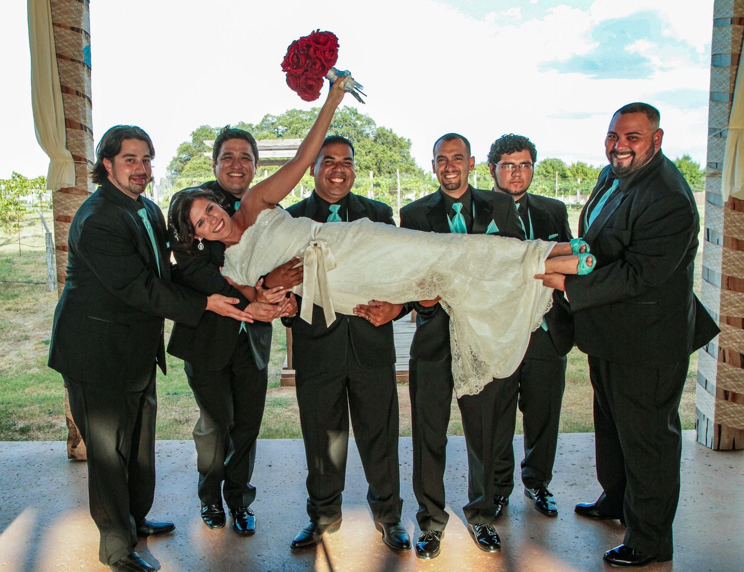 Groom and groomsmen in black suits with teal ties lifting bride in white dress at outdoor wedding celebration.