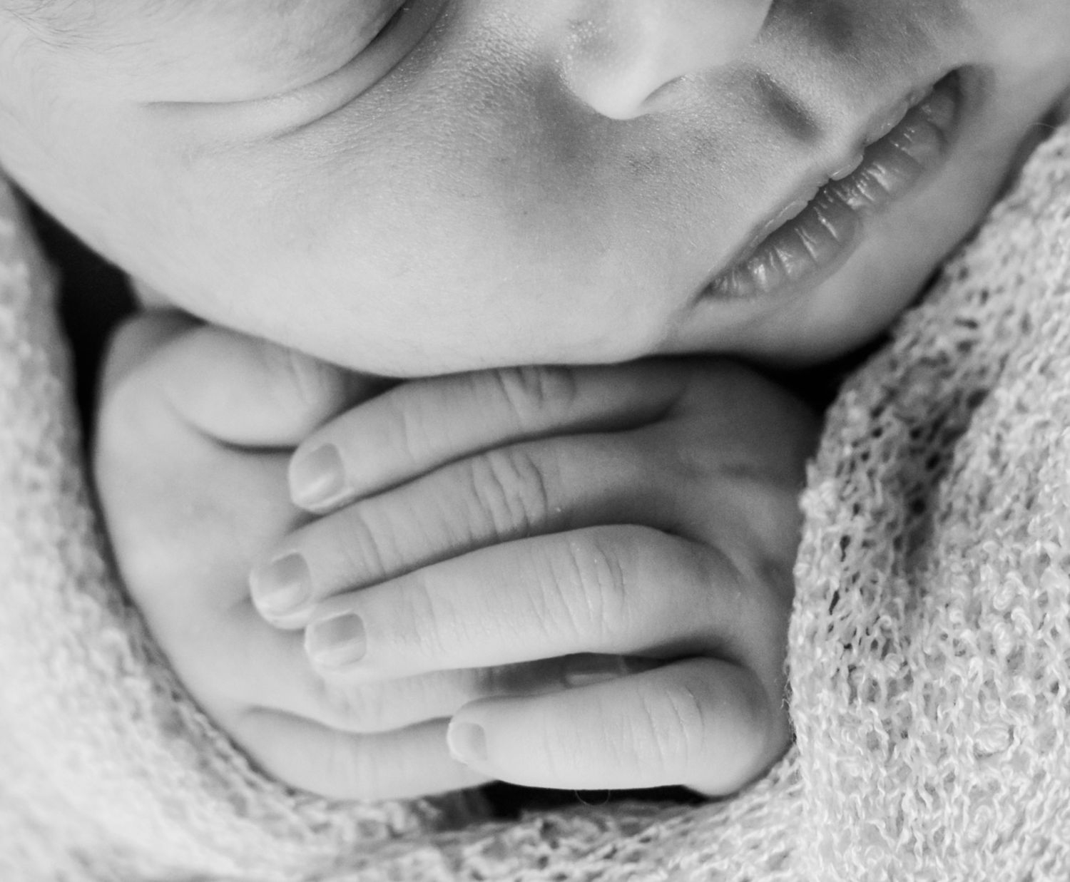Black and white close-up of a sleeping newborn baby with tiny hands tucked under chin, wrapped in soft knit blanket.