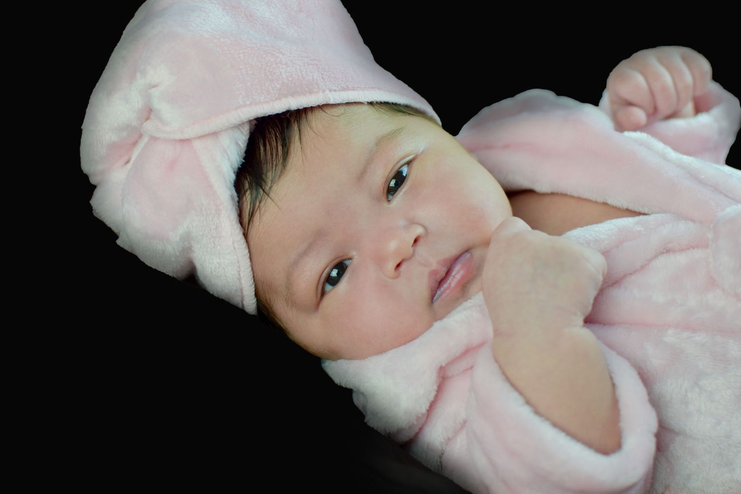 Newborn baby in pink hooded bunny outfit lying on dark background, looking at camera.