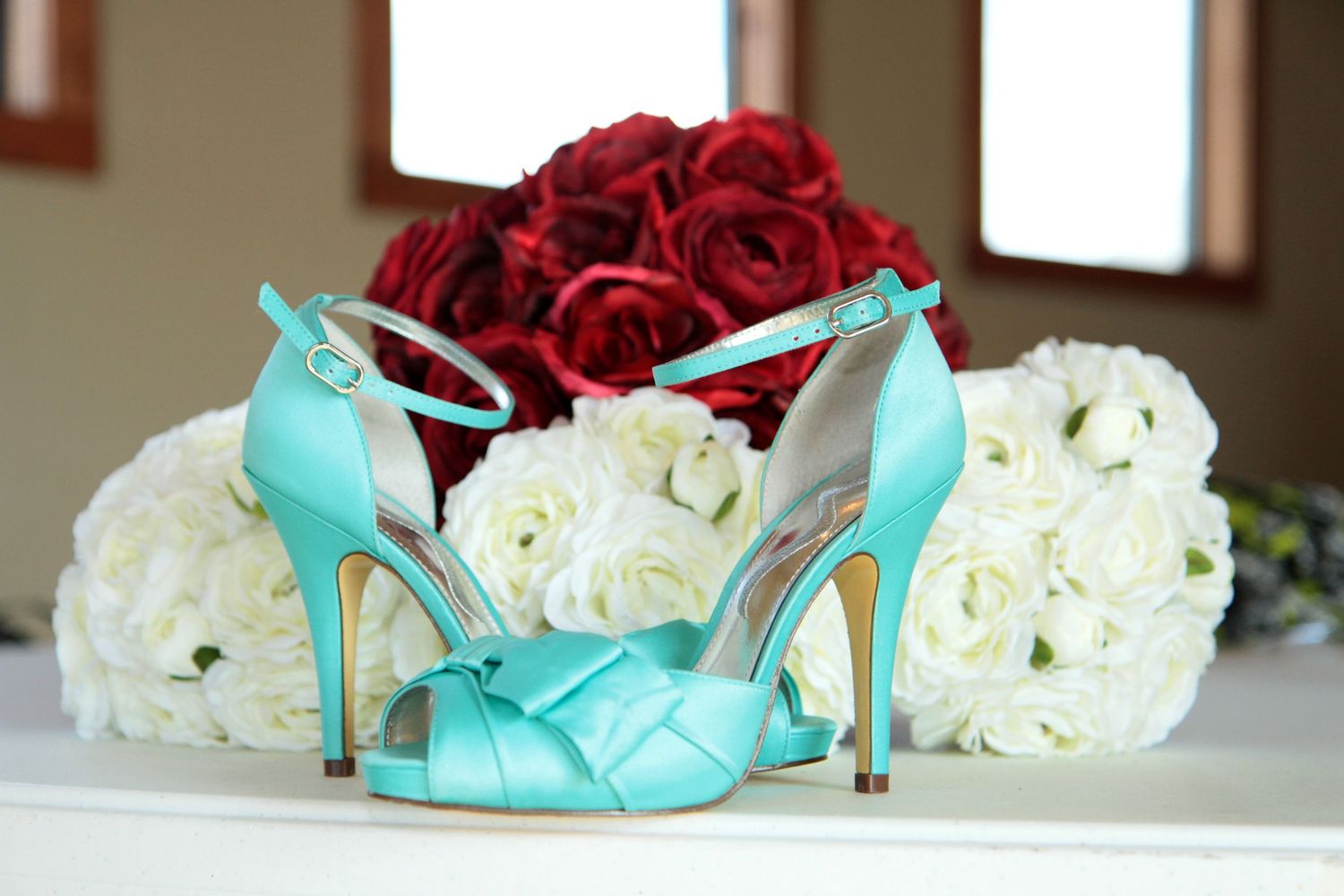 Teal satin high heel sandals with knotted toe strap displayed in front of red and white rose bouquets.