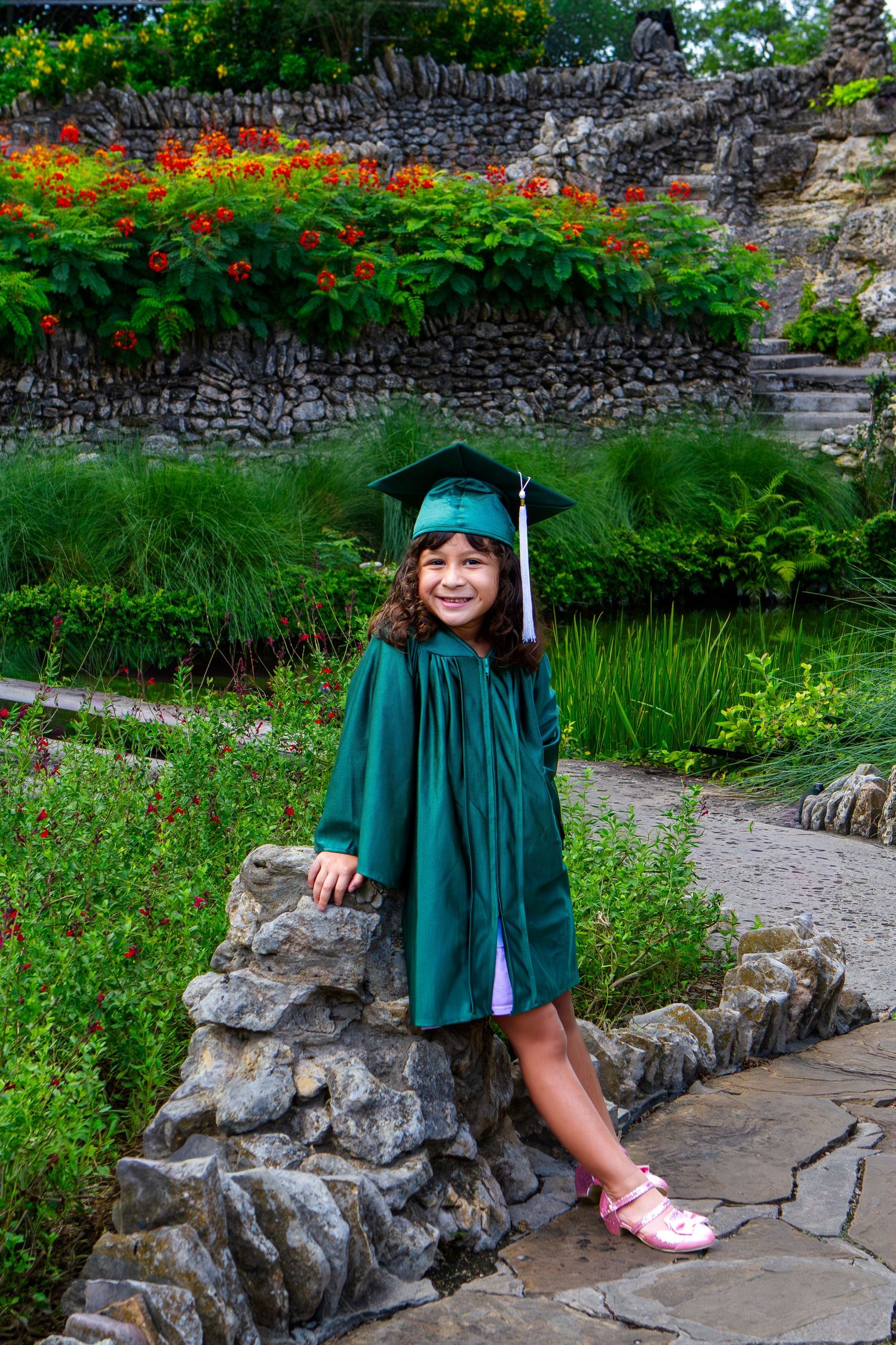 Young girl in green graduation cap and gown smiling in a lush garden with stone walls and red flowers.