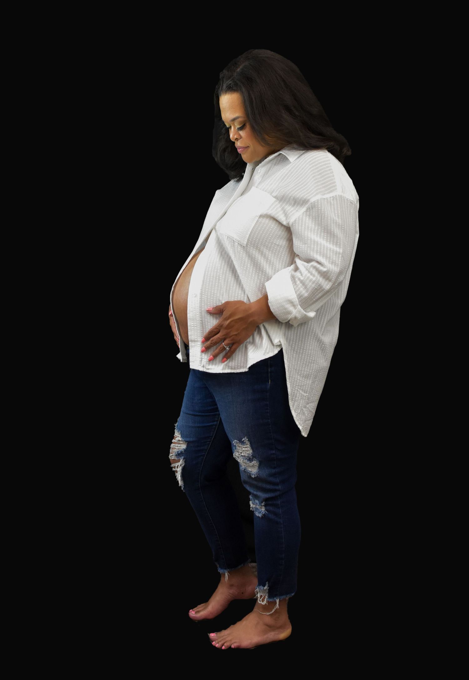 Side view of an expectant mother in a white button-down shirt and distressed jeans standing against a black background in San Antonio, Tx.