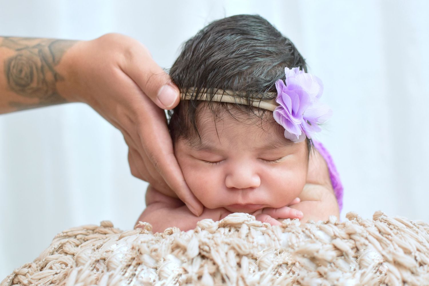 Newborn baby girl sleeping with purple flower headband while adult hand gently supports her head on soft blanket.