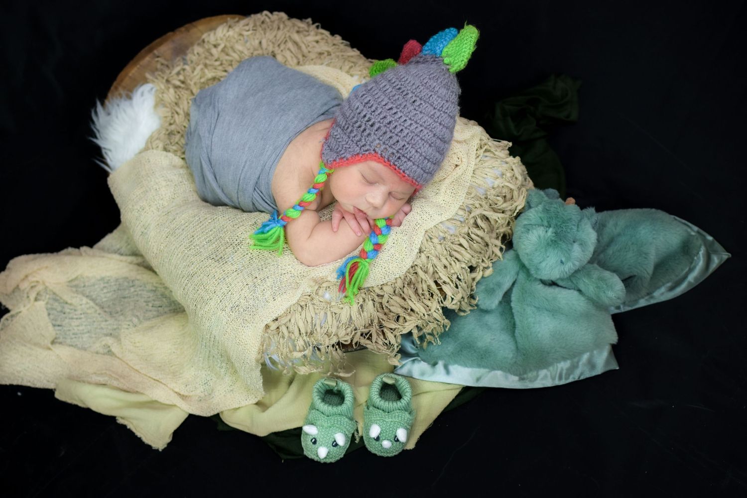Newborn baby sleeping in a nest wearing a gray knit dinosaur hat with colorful spikes and matching green booties.