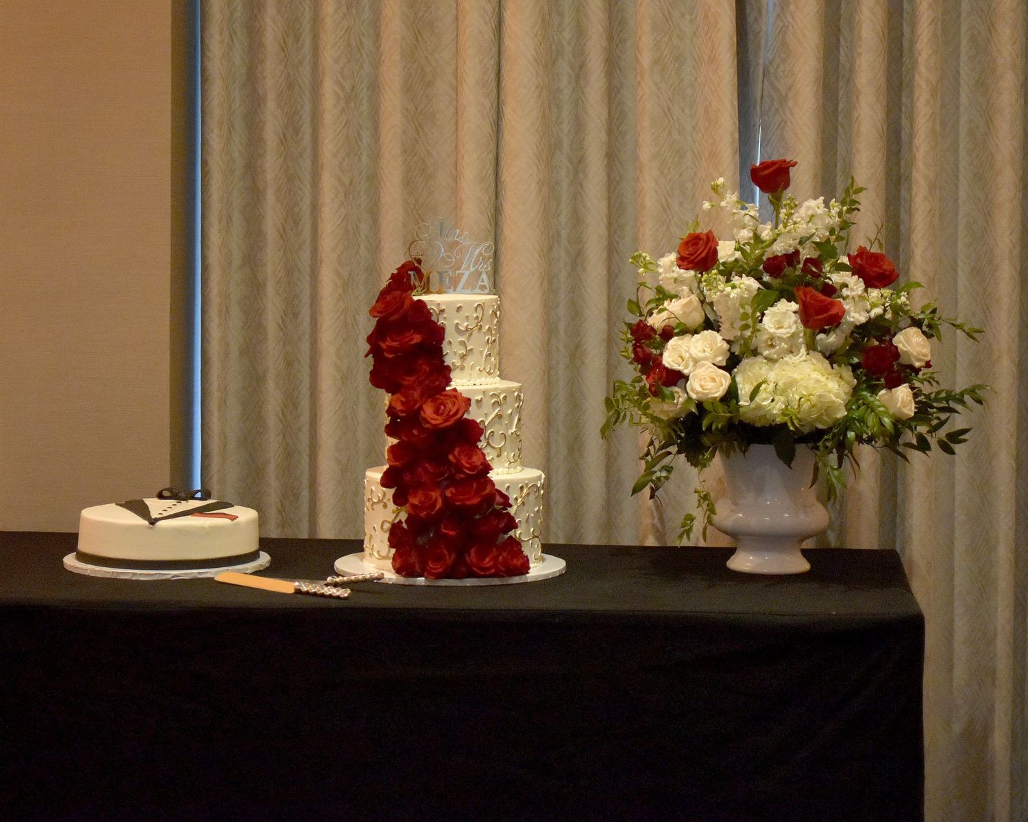 Elegant three-tier white wedding cake adorned with red roses beside a floral arrangement of red and white flowers in a white urn.