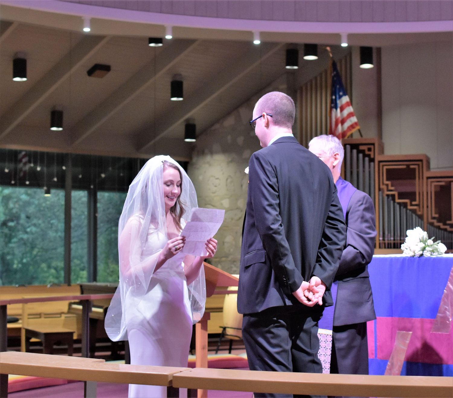 Bride reading vows to groom at church altar during wedding ceremony, officiant standing nearby.