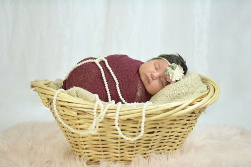 Newborn baby sleeping peacefully in a wicker basket, wrapped in burgundy fabric with pearl beads and floral headband.