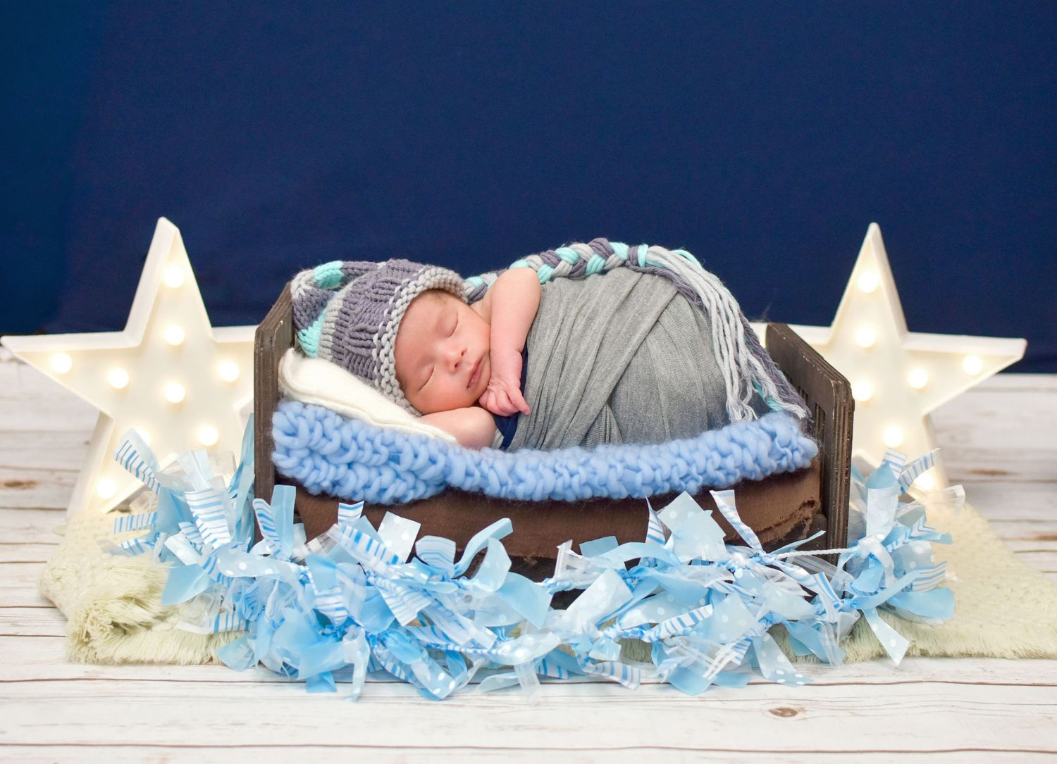 Newborn baby sleeping in a wooden bed with blue blanket, star decorations, and gray knit hat in a studio photo.