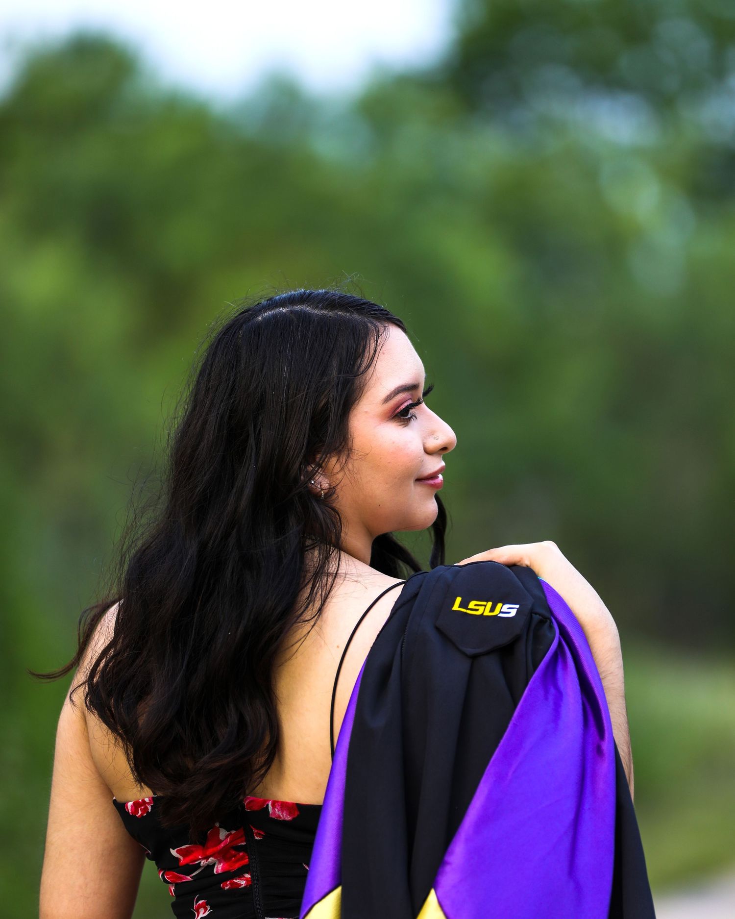 A student in graduation attire with long dark hair stands against a blurred green background showing LSU in her senior graduation photo.