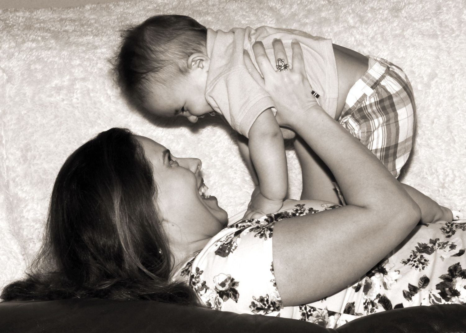 Black and white photo of a mother lying down, joyfully lifting her smiling baby above her face.